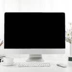 A closeup of a computer, a cup of coffee, a vase of flowers, and more on a white desk, indoors
